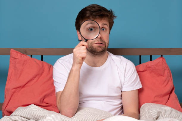 istock Funny young man looking through magnifying glass sitting in the bed 1214909423