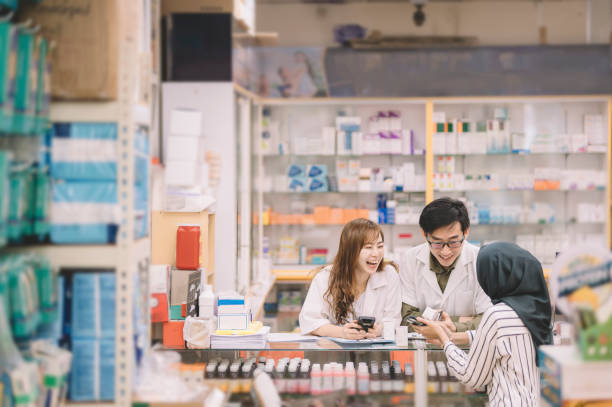 eine asiatische chinesische apothekerin mit labormantel erklärt ihrer weiblichen malaiden-kundin mit hijab an der apothekentheke zusammen mit einem asiatischen chinesischen männlichen apotheker - healthcare and medicine smiling group of people lab coat stock-fotos und bilder