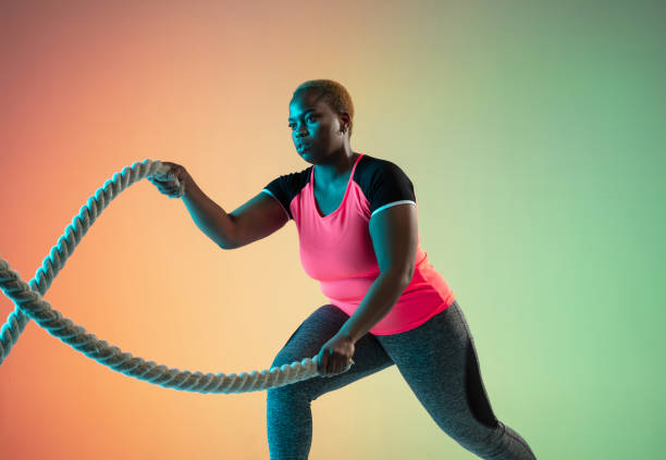 Young african-american plus size female model's training on gradient background Young african-american plus size female model's training on gradient background in neon light. Doing workout exercises with the ropes. Concept of sport, healthy lifestyle, body positive, equality. lift weights stock pictures, royalty-free photos & images