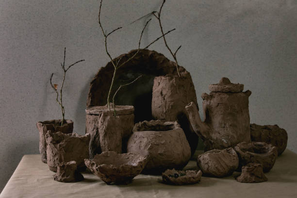 Group of craft handmade raw clay vases and dishes with dry branches in interior on table with linen tablecloth and grey wall behind.