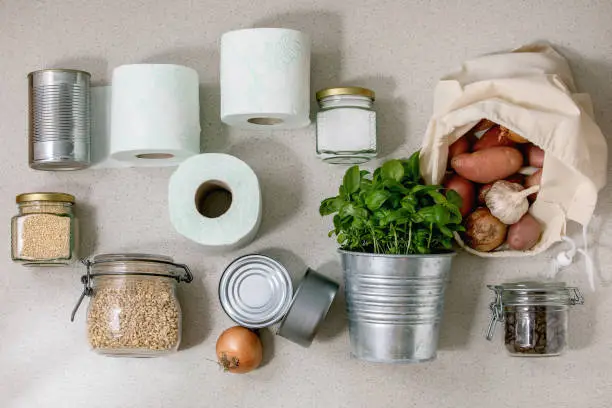 Flat lay of food supplies crisis for quarantine isolation period. Different glass jars with grains, cans of canned food, vegetables, toilet paper, basil.