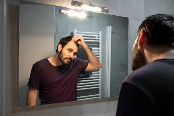 Photo of Handsome young man touching his hair with hand and grooming in bathroom at home. White metrosexual man worried for hair loss and looking at mirror his receding hairline.
