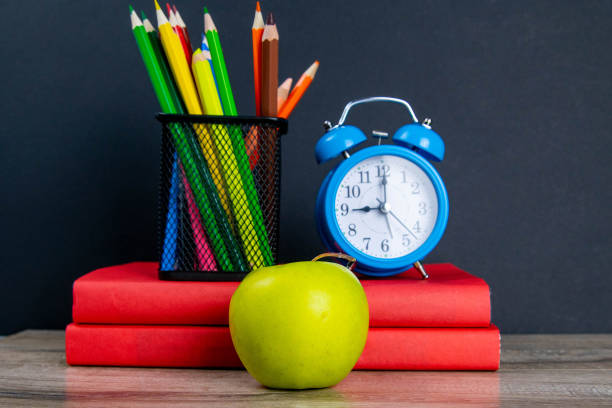 istock The blue alarm clock along with multi-colored pencils stand on a stack of red books, next to an apple 1214932186