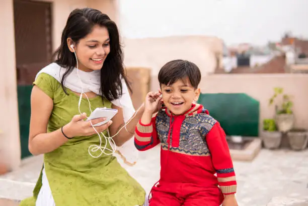 An aunt listening music and sharing headphones with her cute little nephew. They are sitting together and enjoying hair leisure in fresh air at day time. She is wearing traditional Indian dress salwar Kameez and Dupatta.
