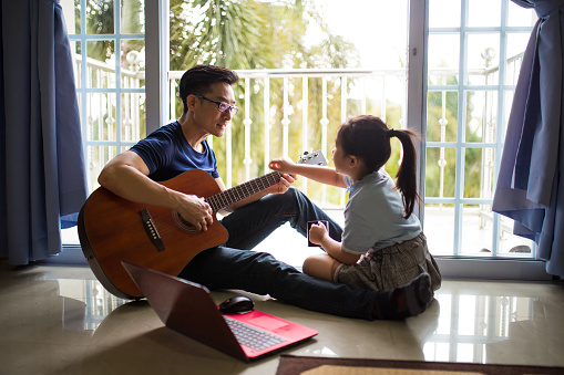 An Asia Chinese businessman work from home. Playing Guitar during coffee break time. Daughter sitting beside and spending wonderful time with him