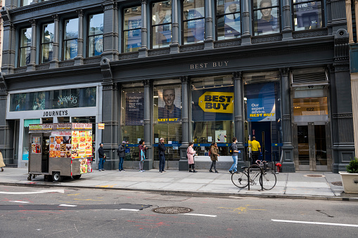 New York City, USA - March 20, 2020: Customers wait in line, spaced a few feet apart, to enter a Best Buy store on Broadway in the Noho neighborhood of Manhattan. Due to the coronavirus pandemic, only a certain number of people are allowed in the store at one time.