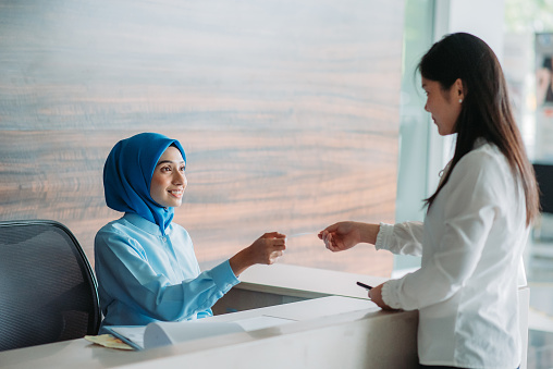 Asian chinese female hijab receptionist getting payment from customer with smiling face at lobby registration counter in hospital