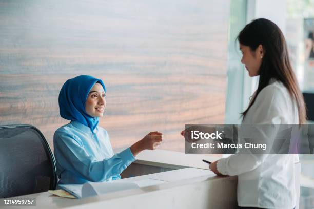 Asian Chinese Female Hijab Receptionist Getting Payment From Customer With Smiling Face At Lobby Registration Counter In Hospital Stock Photo - Download Image Now