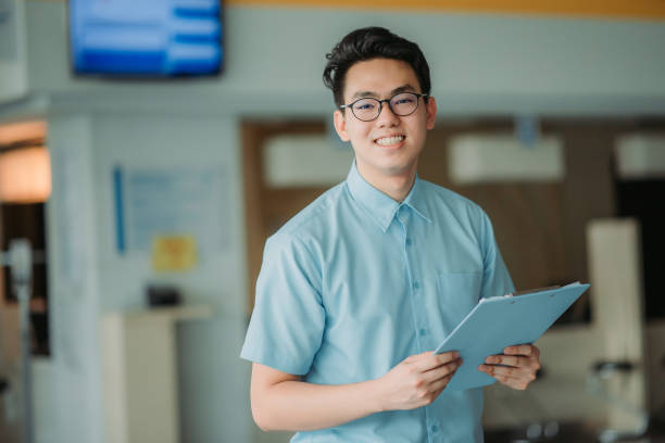 Portrait of chinese male medical healthcare worker hand holding note pad with smiling face standing at entrance lobby of hospital stock photo