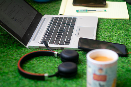 Low shot of laptop on grass with headphones, mobile phone, notepad and a cup of chai tea coffee as someone works from home as a digital nomad freelancer. The artificial grass offers a low maintenance garden in most small homes