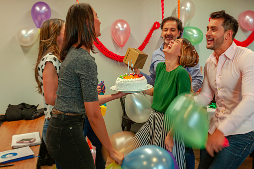 It is party time inside an office in Bogota, Colombia. Colleagues have decorated their place of work and are celebrating with singing, balloons, a cake with candles on it, confetti and dancing, to congratulate their young female colleague. It is a very Latin display of emotions. Horizontal format.