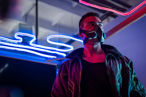 mixed race cyberpunk player in mask standing near neon lighting and looking away