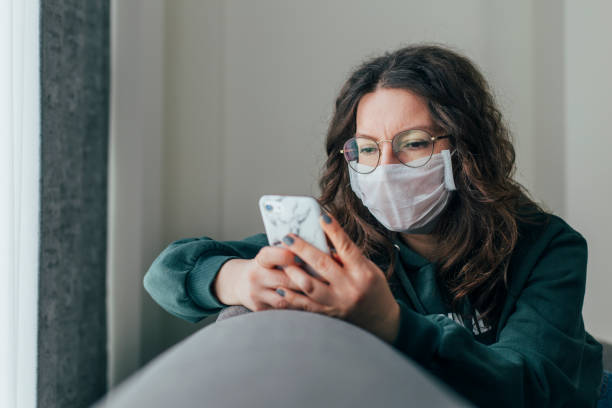 Worried Woman is Reading News on Phone Young woman covering her face with surgical mask and using smartphone to find information about coronavirus spread on internet pandemic illness stock pictures, royalty-free photos & images