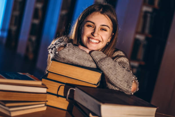 istock Happy smiling female student has completed the exam preparation. The girl leaned on a stack of books sitting in the library at night 1214966610