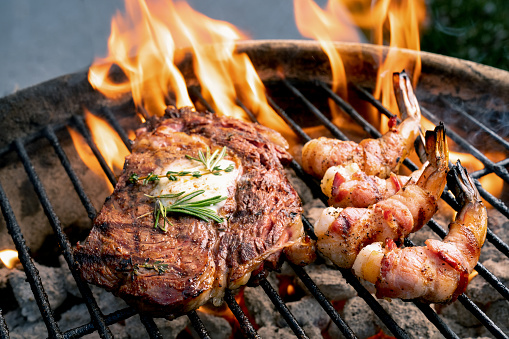 Bacon-wrapped shrimp and a ribeye steak on a barbecue grill with flames