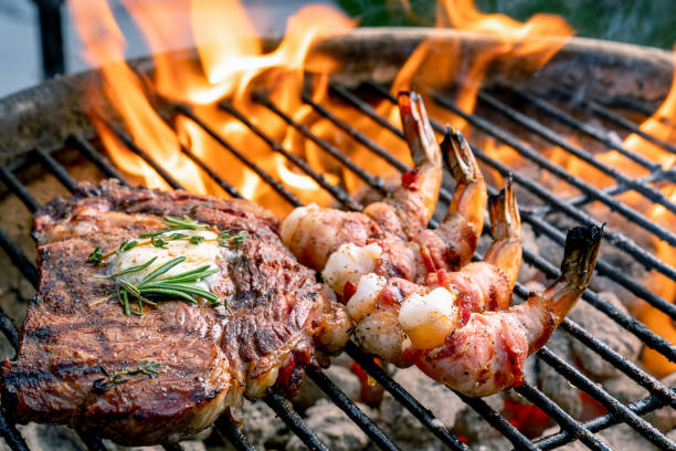 a delicious ribeye steak and bacon wrapped shrimp on a flaming hot grill - surf and turf prepared shrimp seafood steak imagens e fotografias de stock