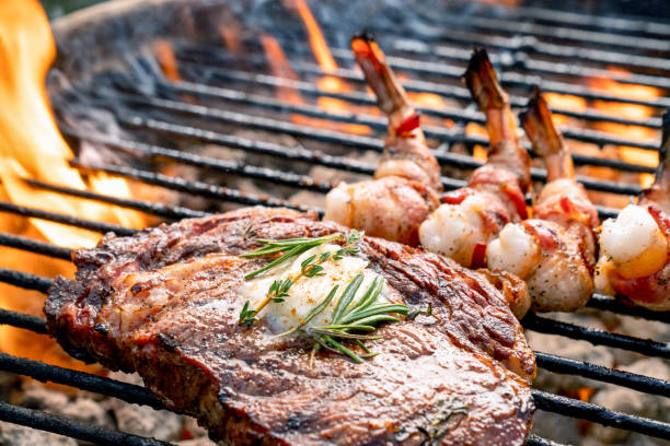 a delicious ribeye steak and bacon wrapped shrimp on a flaming hot grill - surf and turf prepared shrimp seafood steak imagens e fotografias de stock
