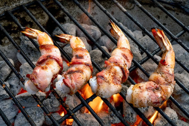 quatre crevettes ou crevettes géantes enveloppées dans du bacon sur un gril au charbon à l’ancienne avec des flammes éclatantes, cuites à un brun doré - surf and turf prepared shrimp seafood steak photos et images de collection