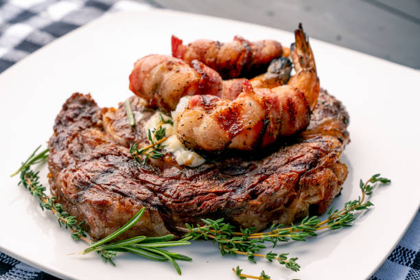 bistecca di ribeye alla brace con timo e rosmarino con gamberi jumbo avvolti in pancetta o gamberi su un piatto, pronti da mangiare - grilled shrimp foto e immagini stock