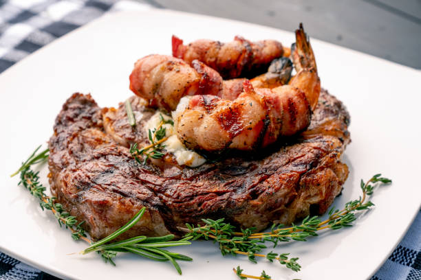 Char-Grilled Ribeye Steak with Thyme and Rosemary with Bacon-Wrapped Jumbo Shrimp or Prawns on a Plate, Ready to Eat Char-Grilled Ribeye Steak with Thyme and Rosemary with Bacon-Wrapped Jumbo Shrimp or Prawns on a Plate, Ready to Eat shrimp seafood stock pictures, royalty-free photos & images