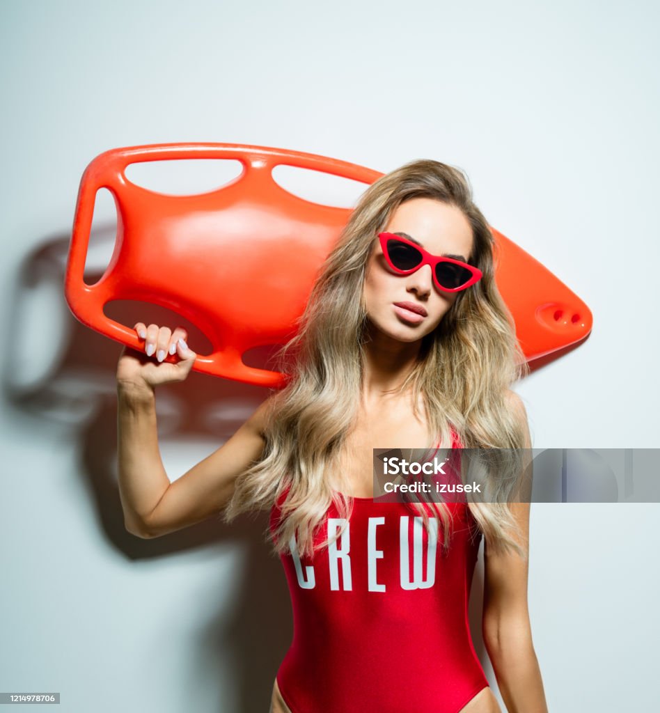 Fashion portrait of young woman holding rescue buoy Summer portrait of beautiful long hair young woman wearing red swimsuit and sunglasses, holding orange rescue buoy on her shoulder, looking at camera. Studio shot on white background. Buoy Stock Photo