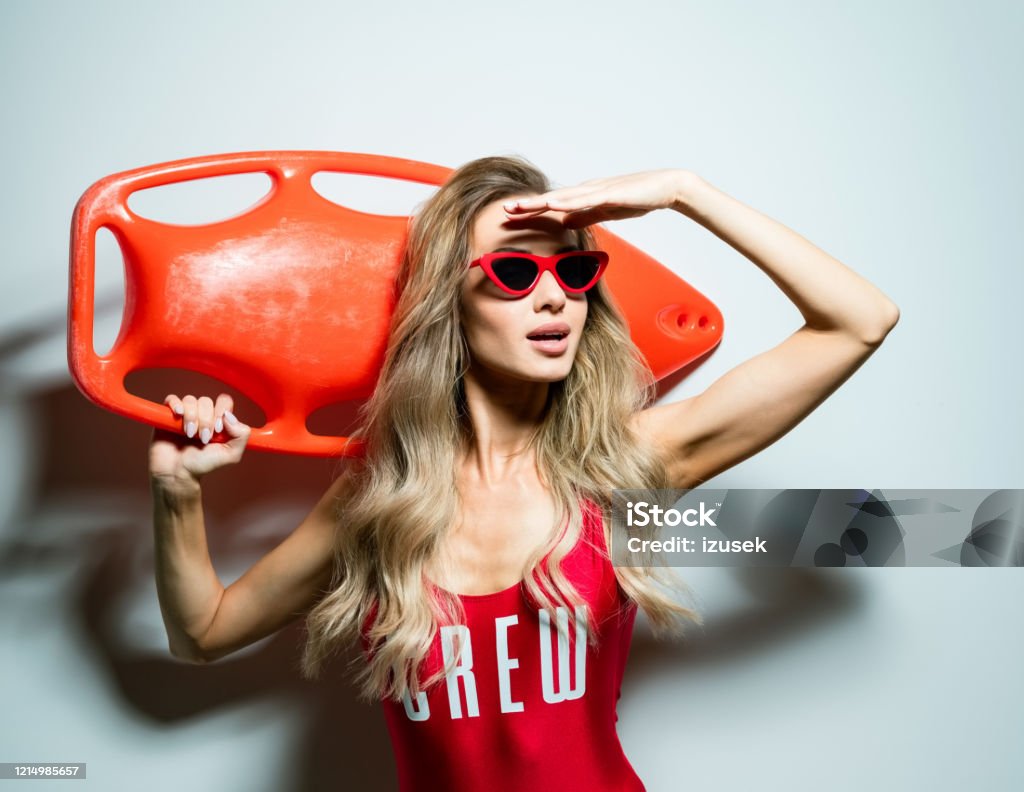 Summer portrait of blonde woman holding rescue buoy Portrait of beautiful long hair young woman wearing red swimsuit and sunglasses, holding orange rescue buoy on her shoulder, looking away. Studio shot on white background. Fashion Stock Photo