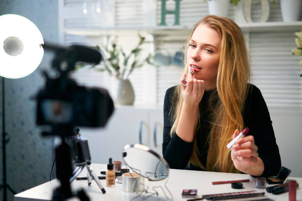 istock Beauty blogger woman filming daily make-up routine tutorial near camera on tripod. Influencer blonde girl live streaming cosmetics product review in home studio. Vlogger female comparing lipsticks 1214987264