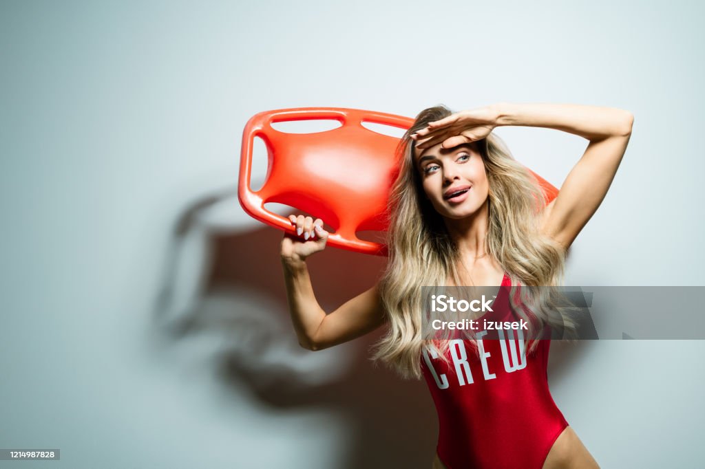 Summer portrait of young woman holding rescue buoy Portrait of beautiful long hair young woman wearing red swimsuit, holding orange rescue buoy on her shoulder, looking away. Studio shot on white background. 25-29 Years Stock Photo