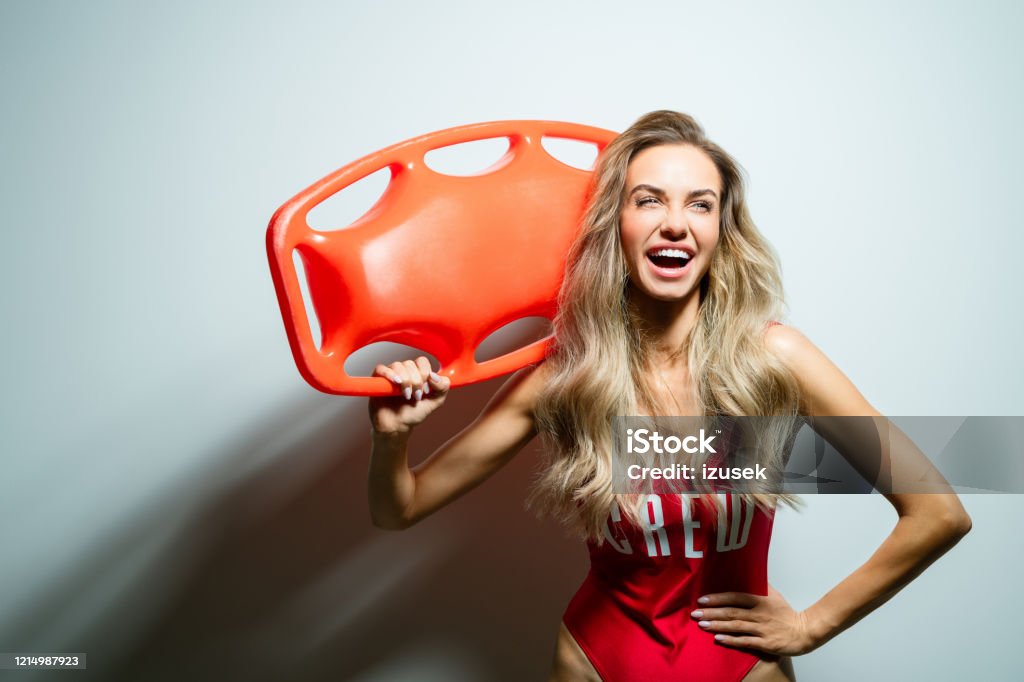 Summer portrait of young woman holding rescue buoy Portrait of happy beautiful long hair young woman wearing red swimsuit, holding orange rescue buoy on her shoulder. Studio shot on white background. Portrait Stock Photo