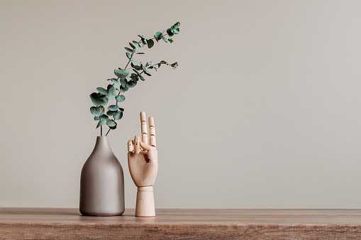 Dry eucalyptus branches in modern vase near wooden hand showing victory sign on the wooden table indoor. Blank space for text. Modern home interior design elements background.