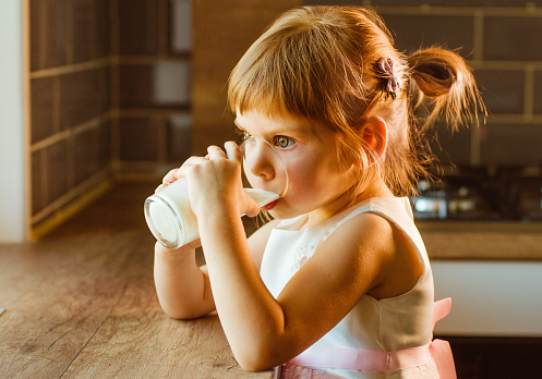 Little girl drinking milk or another dairy product from glass sitting in the kitchen.