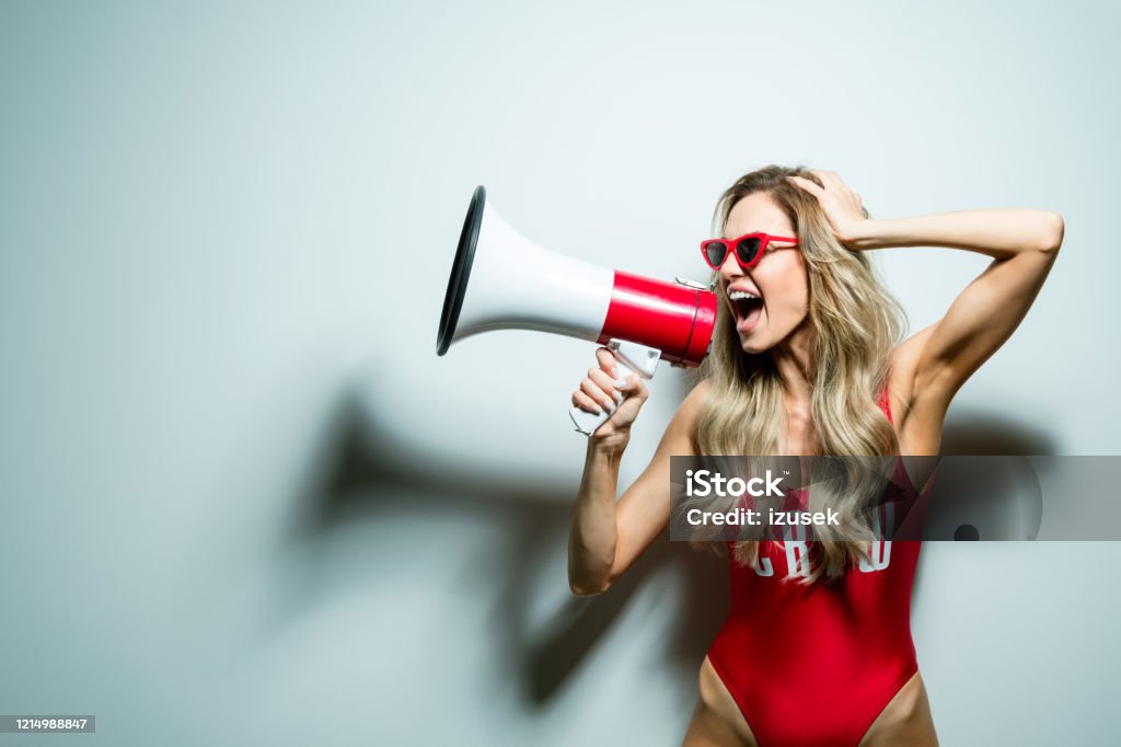 Summer portrait of young woman wearing swimsuit shouting into megaphone Summer portrait of excited blond long hair young woman wearing red swimsuit and sunglasses, holding megaphone. Standing against white background. Studio shot, one person. Megaphone Stock Photo