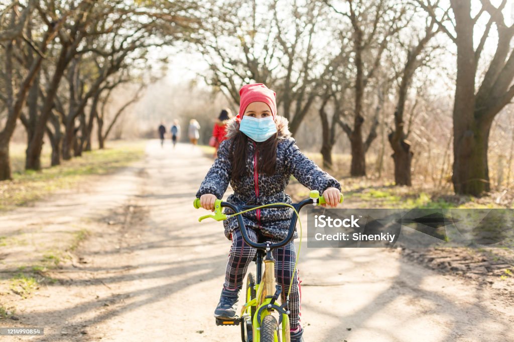 Niña que lleva máscara médica prevenir la gripe, la contaminación y la ovivida-19 montando en bicicleta al aire libre. - Foto de stock de Andar en bicicleta libre de derechos
