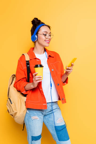 istock cheerful student in headphones using smartphone while holding coffee to go on yellow background 1214995525