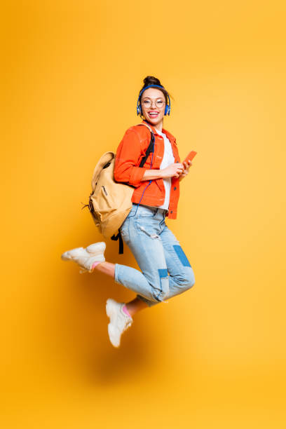 excited student in headset and glasses jumping while holding smartphone on yellow background