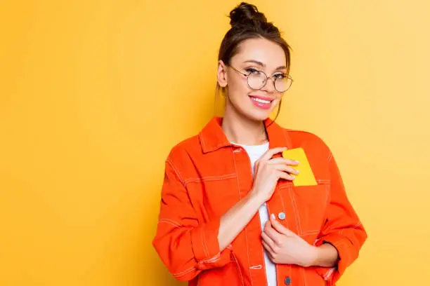 attractive student smiling at camera while taking smartphone out of pocket on yellow background