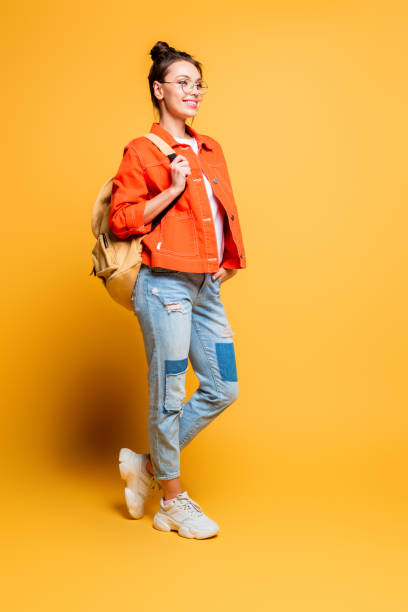 full length view of smiling student with backpack looking away while holding hand in pocket on yellow background