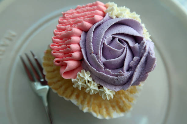 Single iced homemade cup cake on a plate with a fork.  Mauve / lilac / pink icing with a flower (rose) motif. Billericay, Essex, United Kingdom, March 22, 2020.