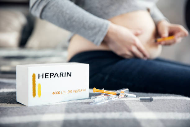 Syringes with medicine and a box with "heparin" inscription lies on the blanket. Pregnant woman makes an injection in the background. Inscriptions on the box made in graphic program. stock photo
