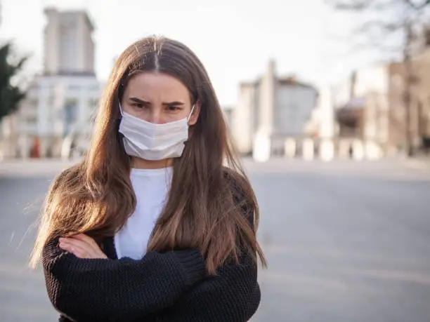 Unhappy cranky frustrated sick girl, brunette woman wearing surgery medical mask with arms crossed feeling bad, ill, being hit by coronavirus, Covid2019 standing outdoors in city looking at you camera