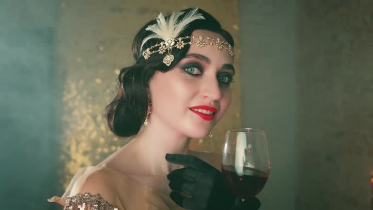 Closeup portrait 20s young beautiful retro woman with glass of red wine in hand. Brunette smile, gold headband decorated bird feathers in vintage luxury dress evening makeup smoky cat eyes bright lips