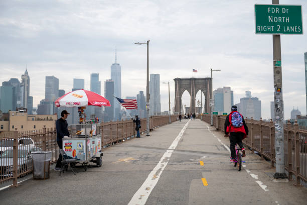 New York City, USA - March 20, 2020: A cyclist rides past a quiet hot dog vendor on the Brooklyn Bridge. A little further down, a man holds an American to encourage, he said, people who were feeling fear during the coronavirus pandemic.