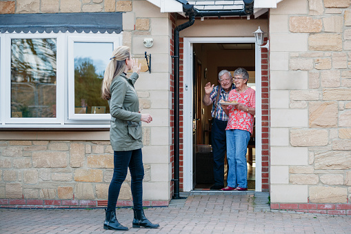 Caucasian female stands back waving from the front door of a bungalow after delivering food, to a senior couple and they wave from their doorway.