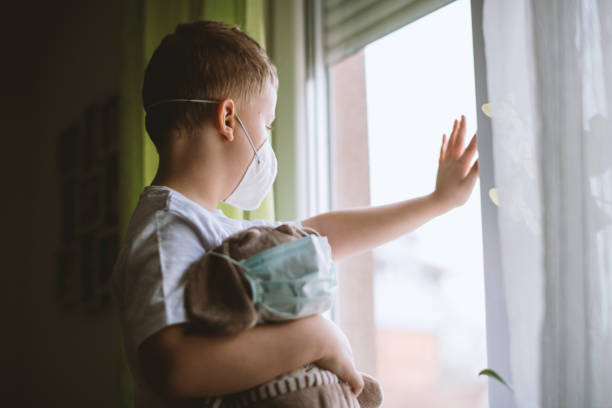 istock Blond hair boy looking through a window wearing protective mask 1215011577