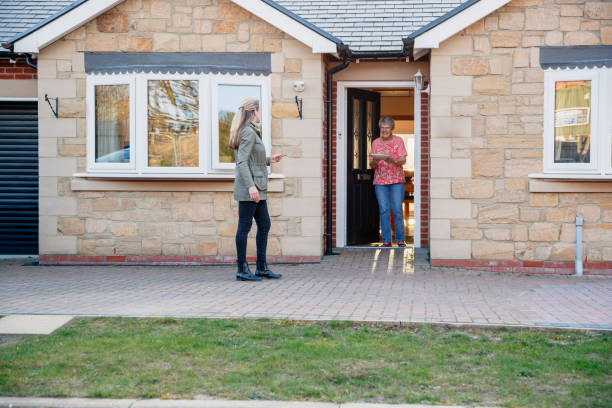 Meals On Wheels Caucasian female stands away from the front door of a bungalow, after delivering a meal to a senior lady who is stood at the front door holding the plate and smiling. meals on wheels photos stock pictures, royalty-free photos & images