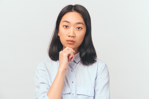Girl holds chin and looks thoughtfully at camera, puzzled. Beautiful young Asian appearance with black hair and brown eyes dressed in striped office shirt stands isolated white background in Studio