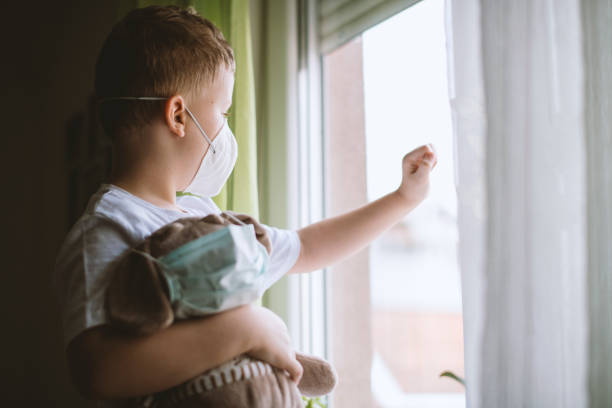 istock Blond hair boy looking through a window wearing protective mask 1215014238