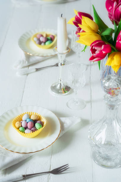 istock Serving Easter table with colorful sweet eggs in cases on the plates, crystal glassware, and fresh tulip flowers on the white wooden table. Family holiday background. Light vertical card. Copy space. 1215018501