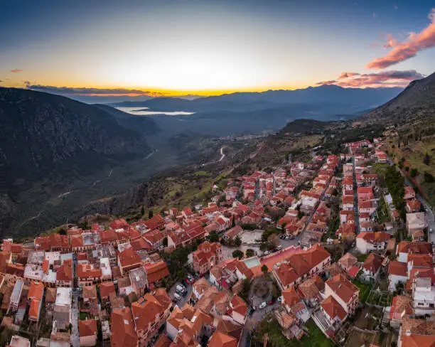 Photo of Aerial view of Delphi, Greece, the Gulf of Corinth, orange color of clouds, mountainside with layered hills beyond with rooftops in foreground