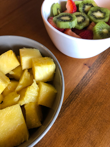 Sliced strawberries and kiwi in a white bowl and chunk pineapple in another displayed on a brown wooden table.